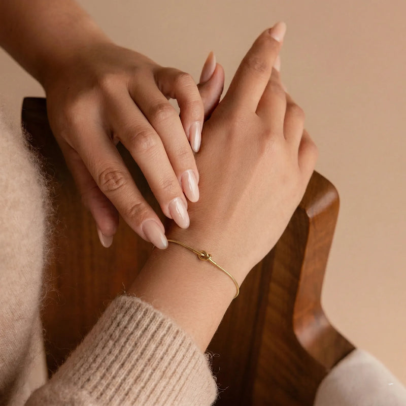 Close-up of a hand wearing a gold bracelet with another hand gently touching it against a warm-toned background.